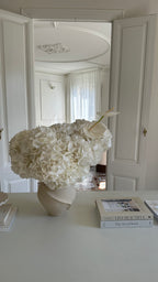 White floral arrangement in a vase on a table with books in a room.