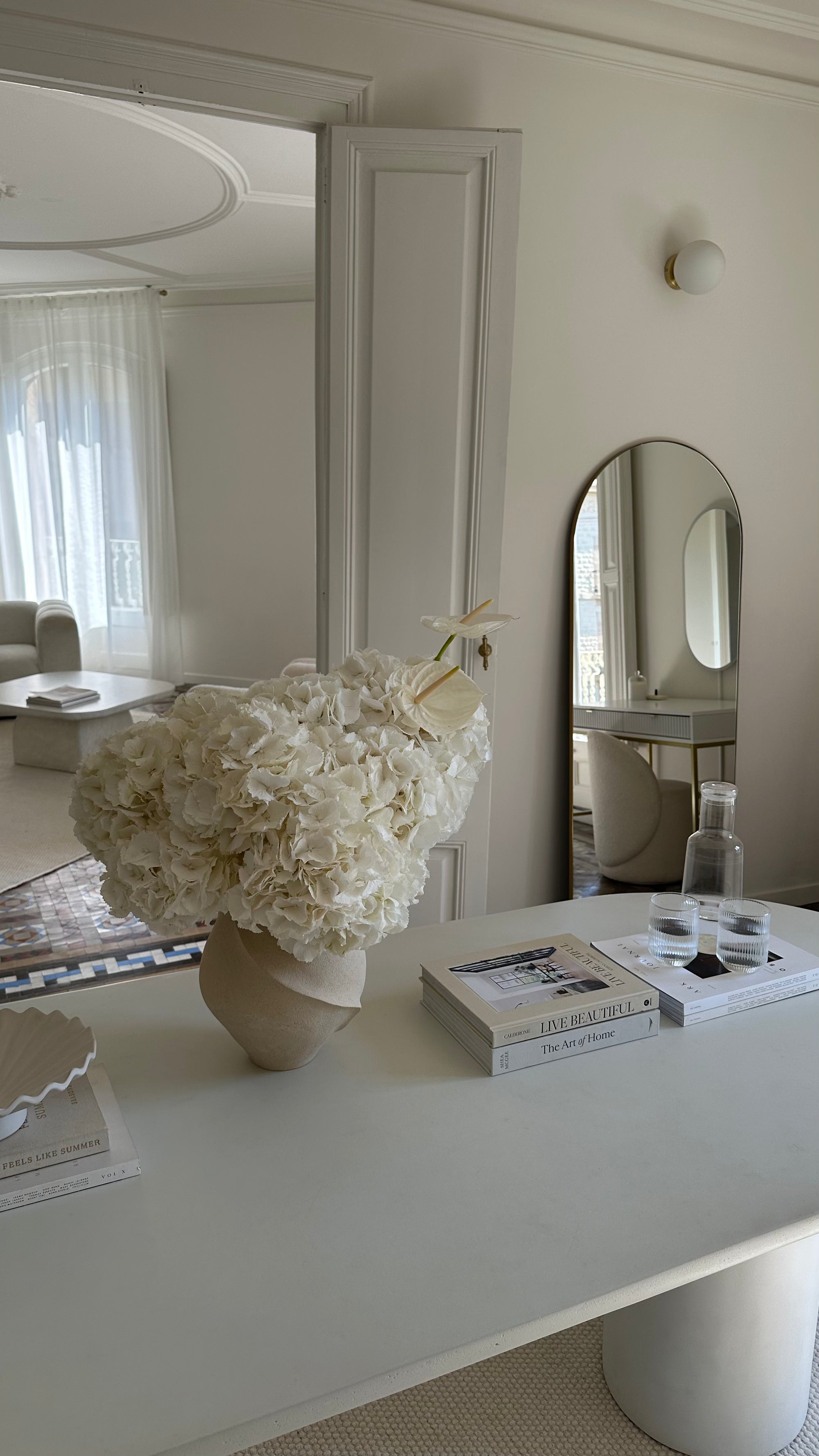 Decorative table with a large white floral arrangement and books in a room with mirrors.